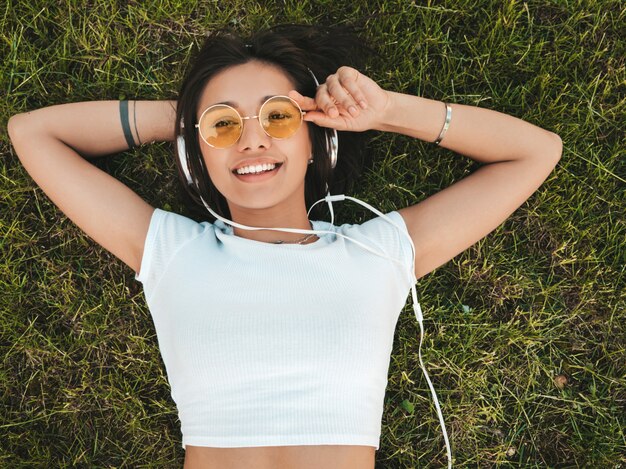 Mode portrait de jeune femme hipster élégant allongé sur l'herbe dans le parc. Fille à la mode tenue à la mode. Modèle souriant profiter de ses week-ends. Femme, Écoute, musique, écouteurs, haut, vue