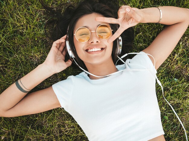 Mode portrait de jeune femme hipster élégant allongé sur l'herbe dans le parc. Fille à la mode tenue à la mode. Modèle souriant profiter de ses week-ends. Femme, Écoute, musique, écouteurs, haut, vue