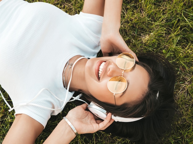 Mode portrait de jeune femme hipster élégant allongé sur l'herbe dans le parc. Fille à la mode tenue à la mode. Modèle souriant profiter de ses week-ends. Femme, Écoute, musique, écouteurs, haut, vue