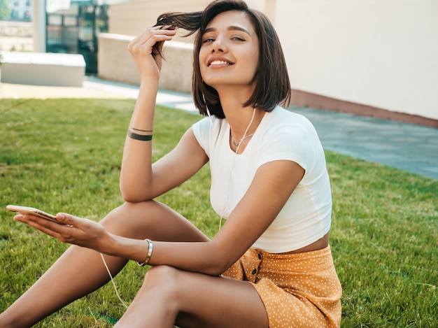 Mode portrait de jeune femme hipster élégant. Fille portant une tenue à la mode mignonne. Modèle souriant profiter de ses week-ends, assis dans le parc Femme écoutant de la musique via des écouteurs