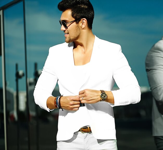 Mode portrait de jeune homme beau modèle sexy en costume blanc décontracté en lunettes de soleil en costume blanc dans la rue