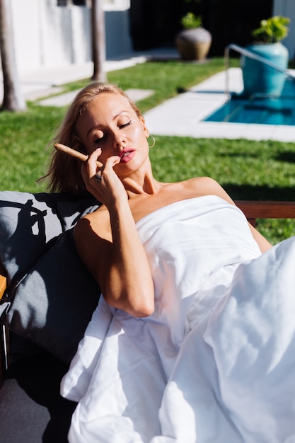 Mode portrait en plein air de femme nue est assise sur un canapé se couvrant avec une couverture tenant un cigare