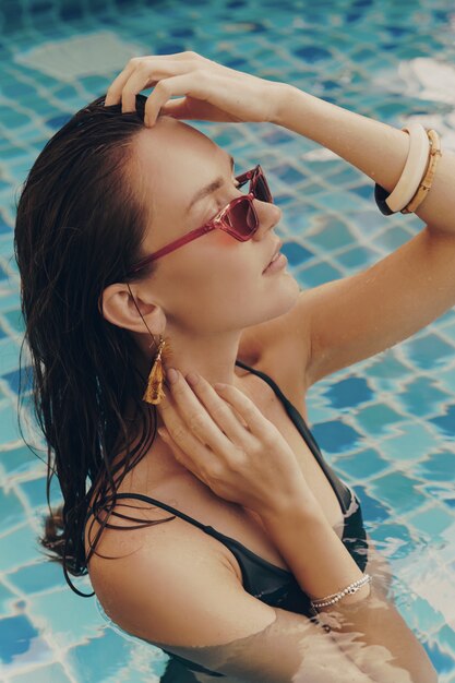 Mode portrait de séduisante femme gracieuse dans des boucles d'oreilles jaunes élégantes avec un corps parfait posant dans la piscine pendant les vacances sur le complexe de luxe.