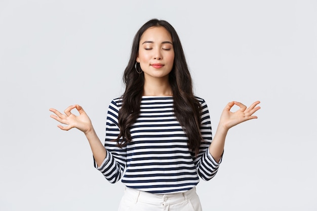 Mode de vie, émotions des gens et concept décontracté. Jeune femme asiatique souriante détendue et patiente aux yeux fermés méditant pour se calmer, faire des exercices de respiration avec les mains dans un geste zen