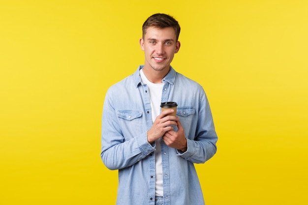 Mode de vie, émotions des gens et concept de loisirs. Beau jeune homme au sourire blanc, commande un café à emporter au café, buvant dans une tasse en papier, ayant une conversation informelle sur fond jaune.