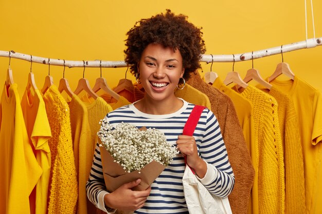 Mode de vie de la femme, mode, concept de consommation. Femme afro-américaine avec l'expression du visage heureux, se tient dans un magasin de vêtements, porte un sac à provisions sur l'épaule, obtient des fleurs de mari, fond jaune