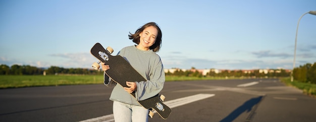 Photo gratuite mode de vie et passe-temps souriante jolie fille asiatique tenant skateboard et marchant vers le soleil sur un vide