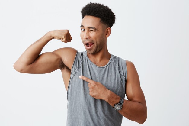 Mode de vie sain. Bouchent le portrait isolé de jeunes hommes sexy à la peau sombre avec des cheveux bouclés en chemise grise sportive montrant le muscle du bras, le pointant, un clin de œil avec la bouche ouverte.