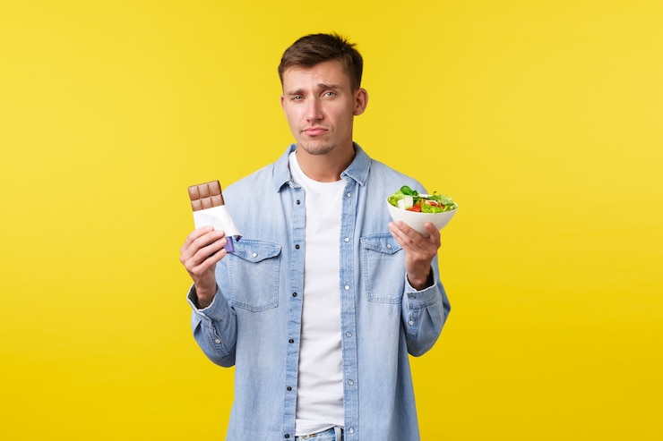 Photo gratuit mode de vie sain et concept d'émotions humaines. beau mec caucasien sombre montrant une salade dans un bol et une barre chocolatée, grimaçant comme réticent à manger des aliments diététiques, debout sur fond jaune