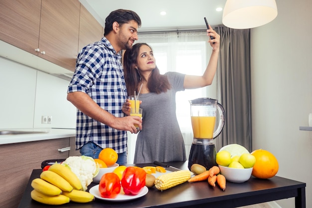 Mode de vie sain et écologique. femme indienne heureuse avec son mari faisant un smoothie dans une grande cuisine, prenant des photos portrait sur l'appareil photo du smartphone.