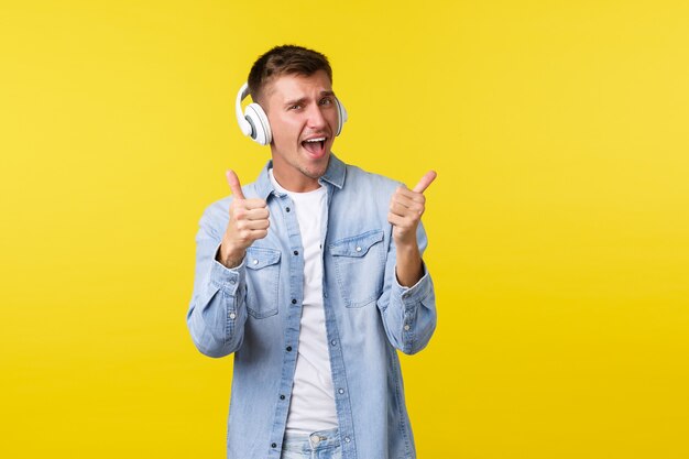 Mode de vie, vacances d'été, concept technologique. Excité bel homme blond heureux vibrant, relaxant avec de la musique géniale, portant des écouteurs et montrant le pouce levé en signe d'approbation, fond jaune.