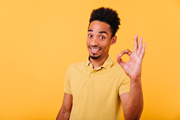 Modèle africain spectaculaire avec coupe de cheveux à la mode posant avec signe correct. Photo intérieure d'un black black étonné en t-shirt d'été.