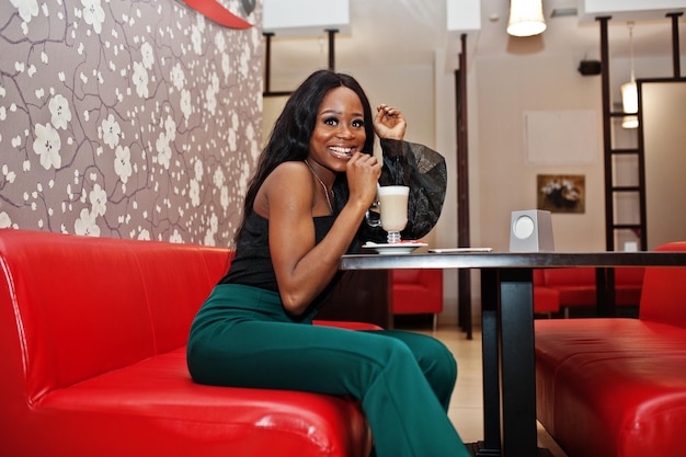 Photo gratuite modèle afro-américain mince de beauté portant un chemisier noir et un pantalon vert à longues jambes buvant du latte au café chinois