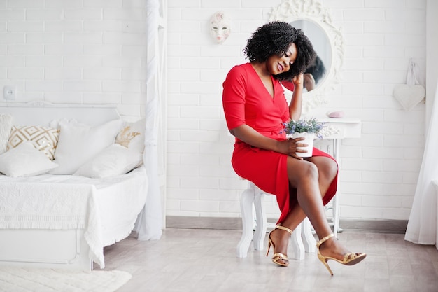 Modèle afro-américain de mode en robe de beauté rouge femme sexy posant robe de soirée assis à une chaise avec pot de lavande contre miroir dans la chambre vintage blanche