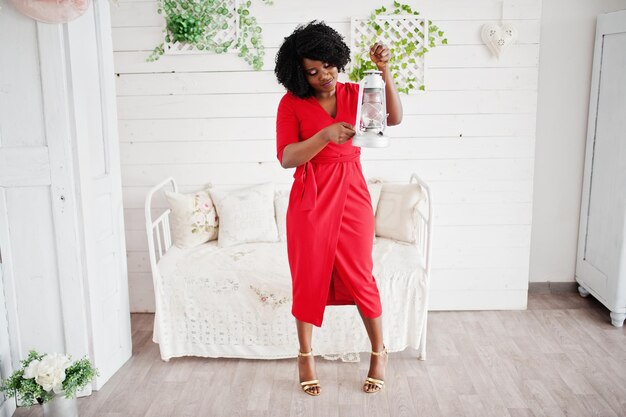 Modèle afro-américain de mode en robe de beauté rouge femme sexy posant une robe de soirée dans une chambre vintage blanche avec une vieille lanterne dans les mains