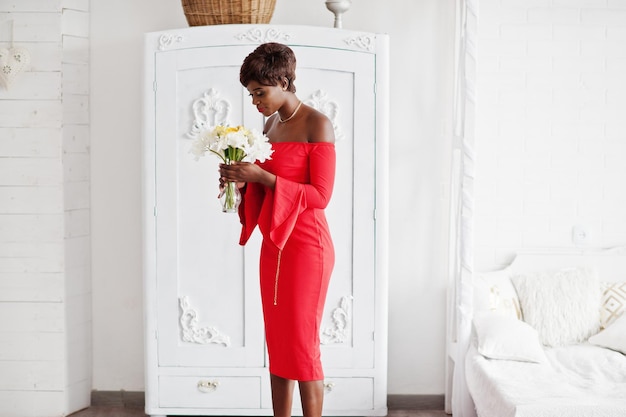 Modèle afro-américain de mode en robe de beauté rouge femme sexy posant robe de soirée tenant des fleurs dans la salle vintage blanche