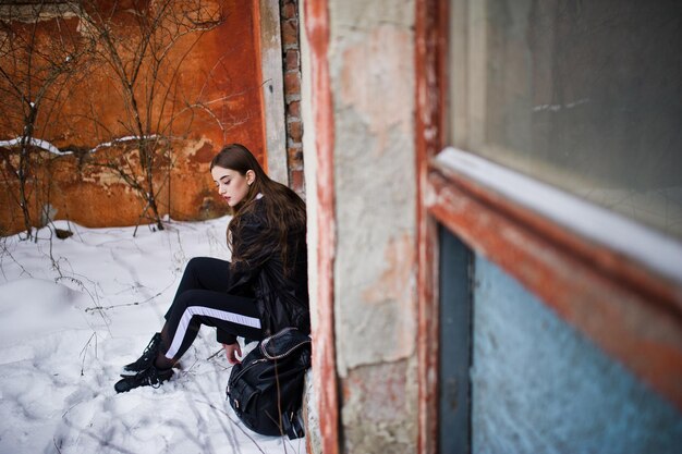 Modèle brune à longues jambes à la mode dans une longue cape noire posée en plein air le jour de l'hiver contre le vieux mur de grunge