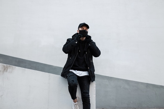 Modèle élégant et beau de jeune homme avec un masque de protection et une casquette noire dans une veste et un sweat à capuche près du mur dans la rue e. style urbain masculin et pandémie