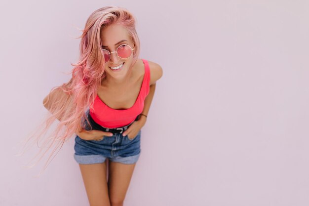 Modèle européen fascinant posant avec les mains dans les poches sur fond blanc Adorable fille bronzée aux cheveux roses souriant pendant la séance photo en intérieur