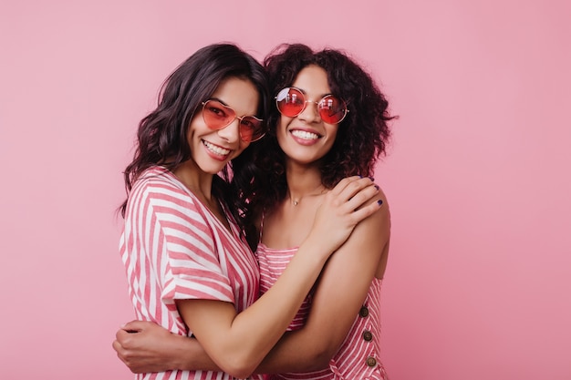 Modèle féminin africain excité en lunettes de soleil rondes embrassant le meilleur ami. Photo intérieure de joyeuse jeune femme noire appréciant