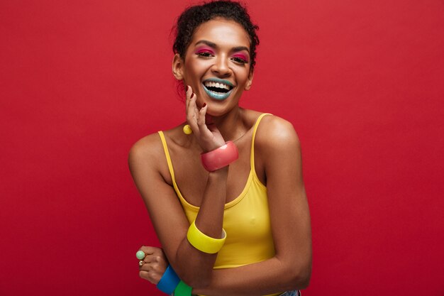 Modèle féminin afro-américain heureux multicolore souriant et se présentant à la caméra en chemise jaune, isolé sur mur rouge