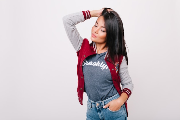 Modèle féminin asiatique de rêve avec des cheveux brillants posant. Portrait intérieur d'une jeune fille brune romantique à la peau légèrement bronzée posant en jeans et t-shirt gris.