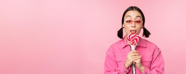 Modèle féminin asiatique stupide et mignon léchant lolipop mangeant des bonbons doux et souriant à la recherche excitée debout sur fond rose