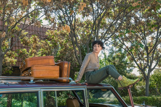 Modèle féminin assis sur une voiture posant pour une séance photo avec des suticases sur le côté