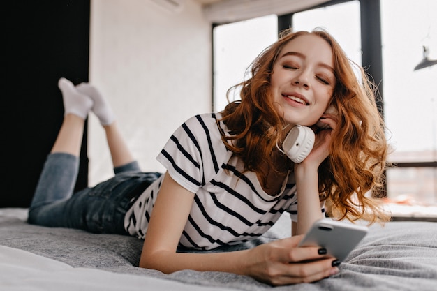 Modèle féminin attrayant avec téléphone à la main allongé sur le lit. Bonne fille détendue appréciant la musique dans les écouteurs.