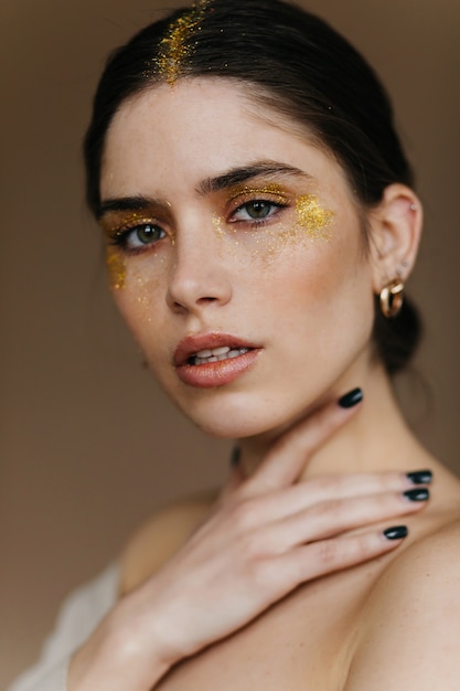 Modèle féminin brune calme posant dans des accessoires dorés. fille aux cheveux noirs à la mode avec du maquillage de fête.