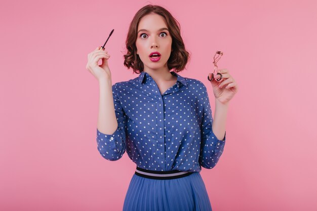 Modèle féminin choqué en tenue bleue posant avec du mascara à la main. belle femme étonnée recourbe ses cils.