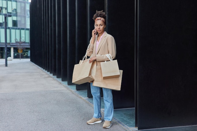 modèle féminin dans des vêtements à la mode porte de nombreux sacs en papier revient du centre commercial après avoir fait des achats réussis fait des poses d'appels téléphoniques en ville attend quelqu'un