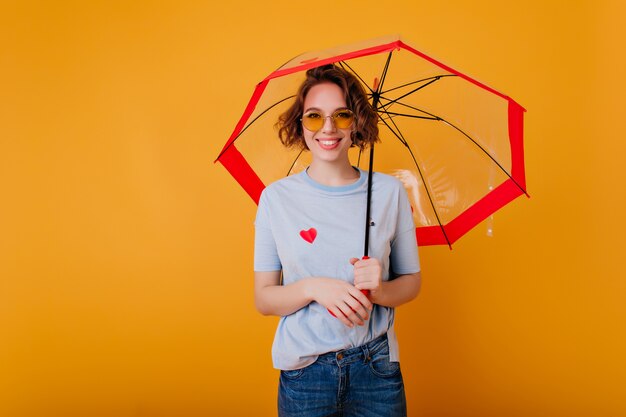 Modèle féminin enthousiaste dans des verres à la mode debout avec un parapluie et souriant. Studio photo de rire fille européenne bouclés avec parasol isolé sur un mur lumineux.