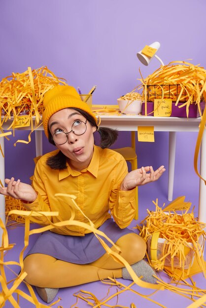 Modèle féminin hésitant en chemise de chapeau jaune et jupe pose les jambes croisées près du bureau écarte les paumes n'a aucune idée ou idée de ce qu'il faut faire en premier est assis sur le sol dans son propre cabinet entouré de déchets de papier py.