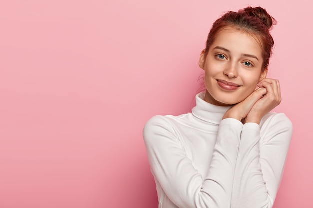 Le modèle féminin heureux incline la tête et sourit doucement, maintient les paumes pressées ensemble près du visage, n'a pas de maquillage, porte un col roulé blanc, regarde directement la caméra avec des yeux bleus, isolé sur fond rose