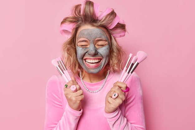 Un modèle féminin joyeux rit avec joie tient des pinceaux cosmétiques pour appliquer un masque d'argile de beauté sourit largement fait une coiffure avec des rouleaux porte une robe de fête et un collier isolés sur fond rose
