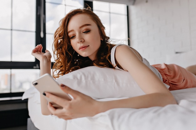 Modèle féminin mignon avec un maquillage sombre allongé sur le lit. Fille de gingembre insouciante regardant l'écran du téléphone.