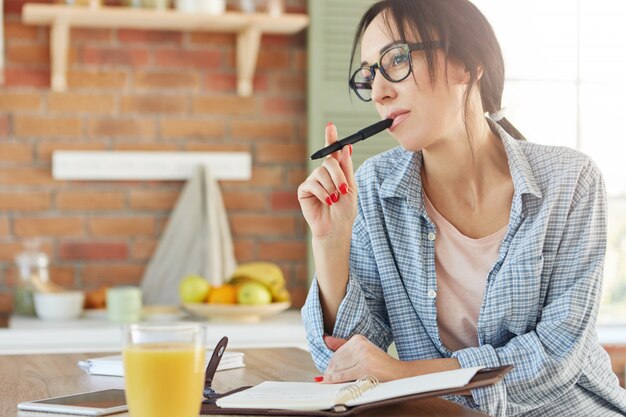 Le modèle féminin pensif garde un stylo, écrit la recette dans le bloc-notes, essaie de se souvenir de tous les ingrédients nécessaires