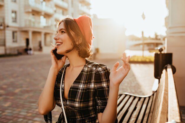 Modèle féminin positif dans des vêtements élégants profitant de la journée d'automne. Photo extérieure d'une superbe femme bouclée en béret rouge parlant au téléphone.