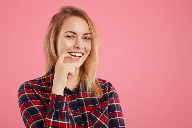 Modèle féminin positif mord le doigt, regarde avec une expression heureuse à la caméra, heureux de rencontrer un beau mâle