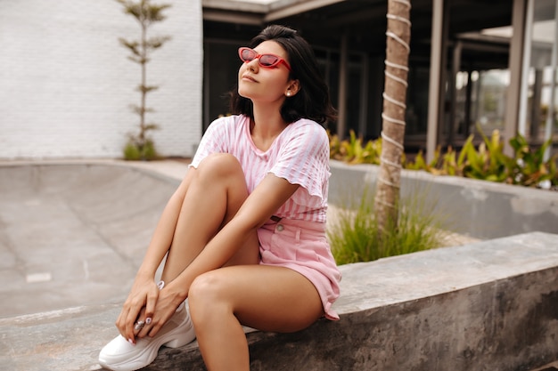 Modèle féminin de rêve en tenue d'été assis sur fond urbain. Tir extérieur d'une femme incroyable en t-shirt rose et lunettes de soleil.