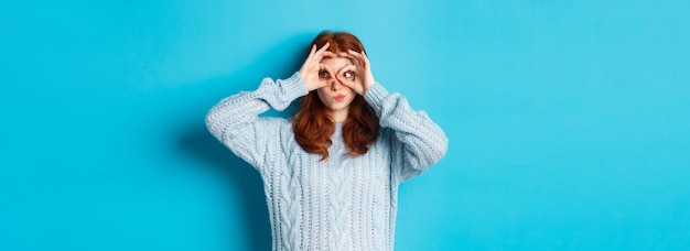 Modèle féminin rousse drôle en chandail regardant la caméra à travers des lunettes de doigts exprimer son intérêt et