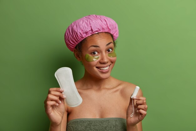 Modèle féminin souriant en bonne santé enveloppé dans une serviette de bain, porte des patchs de beauté verts sous les yeux