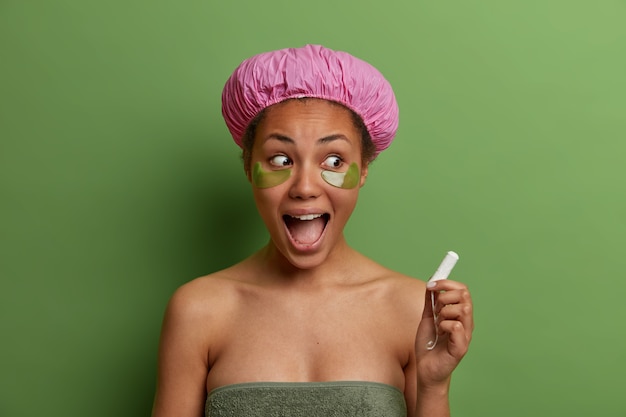 Modèle féminin souriant en bonne santé enveloppé dans une serviette de bain, porte des patchs de beauté verts sous les yeux