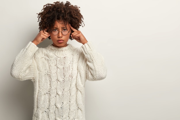 Le modèle féminin stressant garde les doigts sur les tempes, a une expression boudeuse, a l'air affligé, a la migraine après un travail fatigué, réfléchit fort, vêtu d'un pull en tricot blanc, se tient debout.