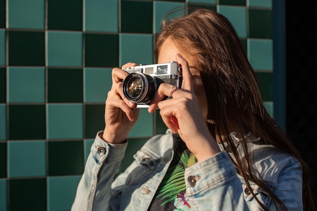 Modèle de fille cool avec caméra rétro portant une veste en jean, cheveux noirs à l'extérieur sur le mur de la ville dans un fond de cage. S'amuser dans la ville avec un appareil photo, photo de voyage du photographe.
