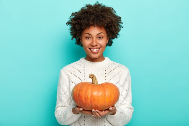 Modèle heureux avec des cheveux afro, détient une grosse citrouille orange mûre, connaît une bonne recette pour préparer un délicieux repas bio, porte un pull blanc