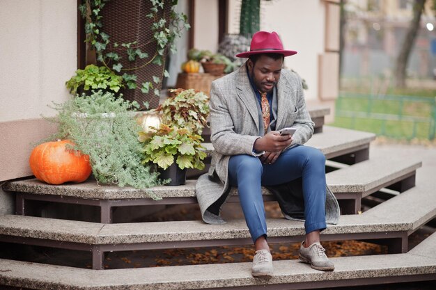 Modèle d'homme afro-américain élégant en manteau gris veste cravate et chapeau rouge assis contre des citrouilles Humeur d'automne