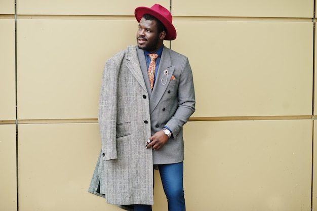 Photo gratuite modèle d'homme afro-américain élégant en manteau gris veste cravate et chapeau rouge posé sur fond doré