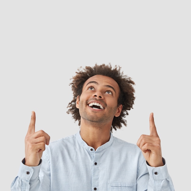 Photo gratuite le modèle homme positif regarde joyeusement vers le haut, a un sourire amical, montre des dents blanches parfaites, indique avec les deux doigts antérieurs au-dessus de la tête